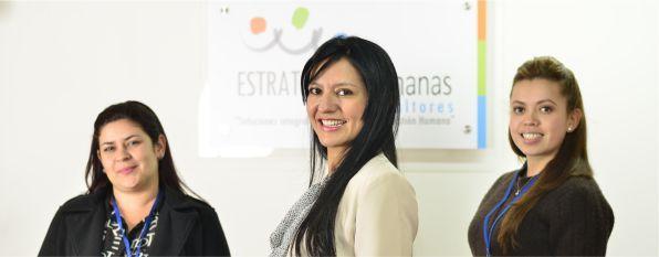 Three women smiling and standing in front of a company sign in an office setting.